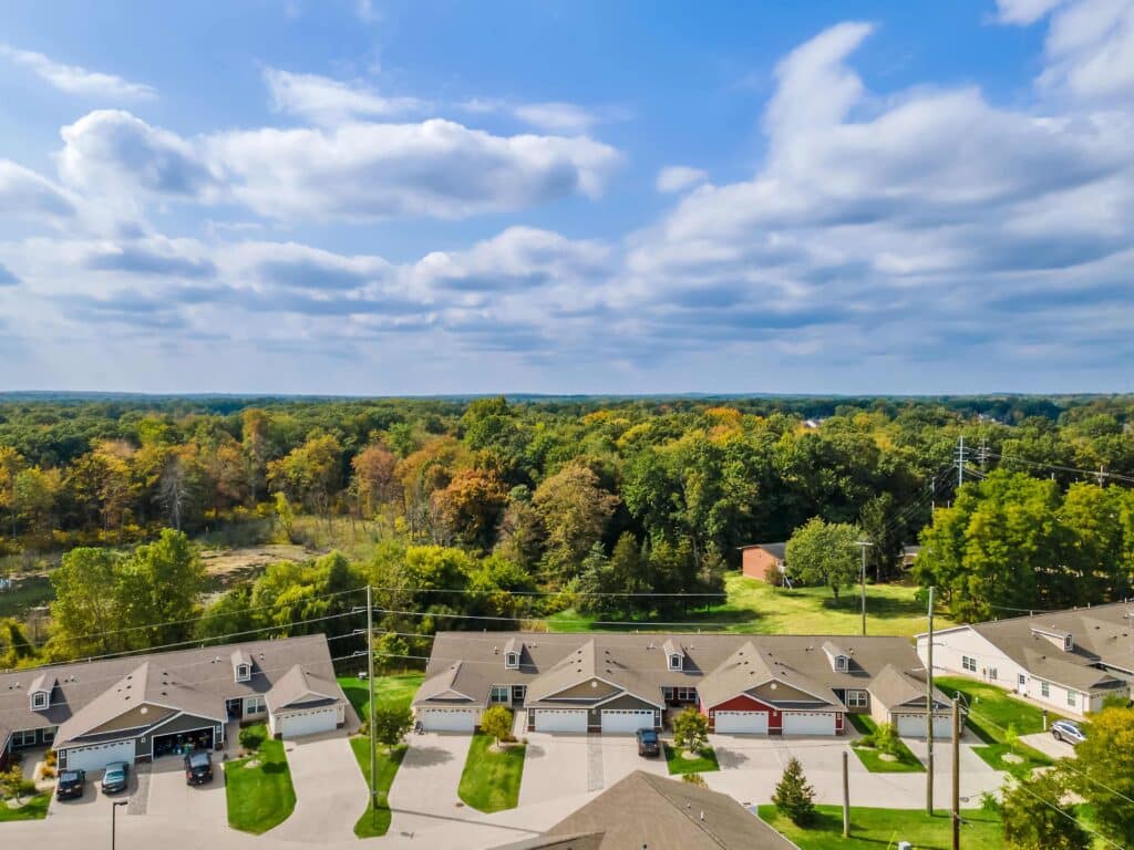 Aerial view of a row of suburban houses with driveways, cars parked outside, and a wooded area in the background under a partly cloudy sky.