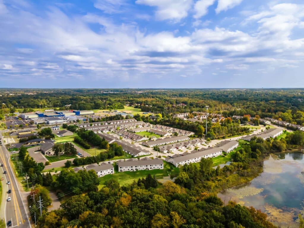 Aerial view of a suburban neighborhood with rows of houses, surrounding trees, a pond, and commercial buildings in the background under a partly cloudy sky.