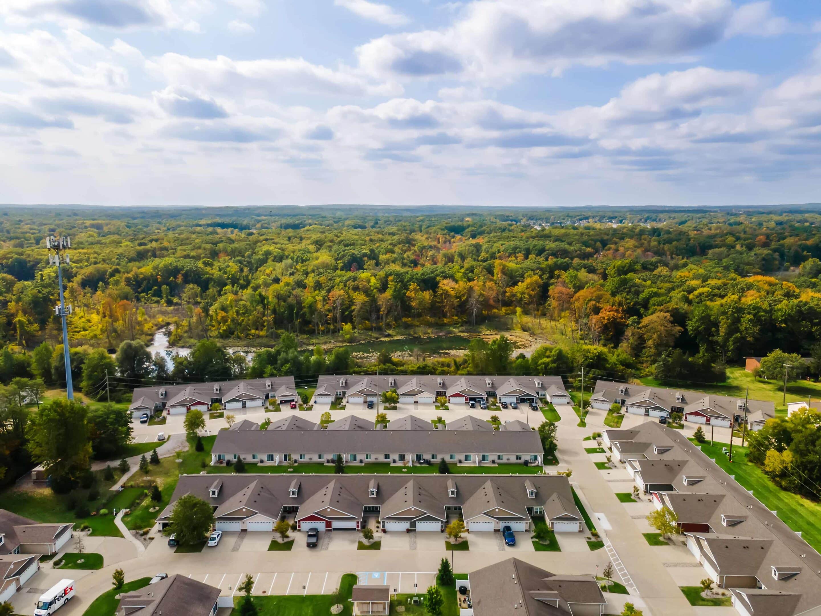 Aerial view of a suburban neighborhood with rows of houses, surrounding trees, parked cars, and a forested area in the background under a partly cloudy sky.
