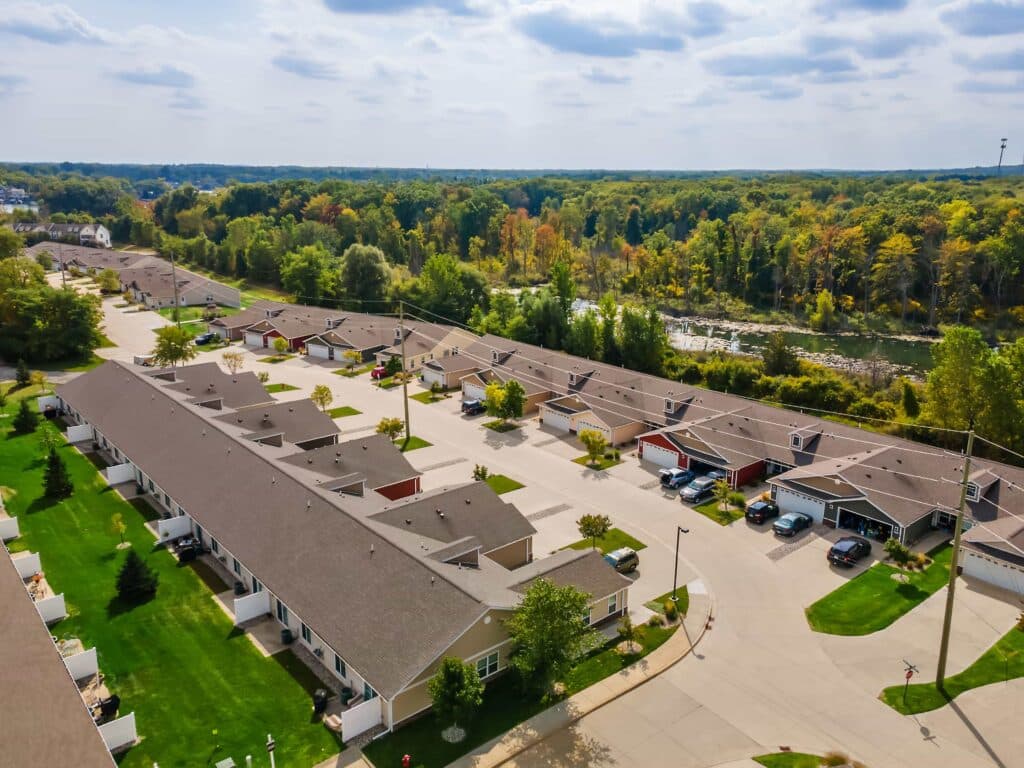 Aerial view of a residential neighborhood with rows of townhouses, green lawns, parked cars, and trees in the background under a partly cloudy sky.
