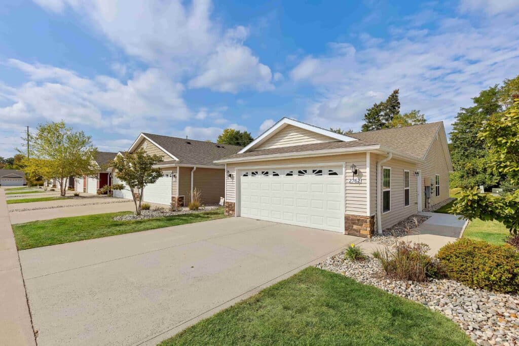 Single-story, beige suburban house with a two-car garage and driveway, surrounded by green lawn, small trees, and landscaping, under a partly cloudy sky.