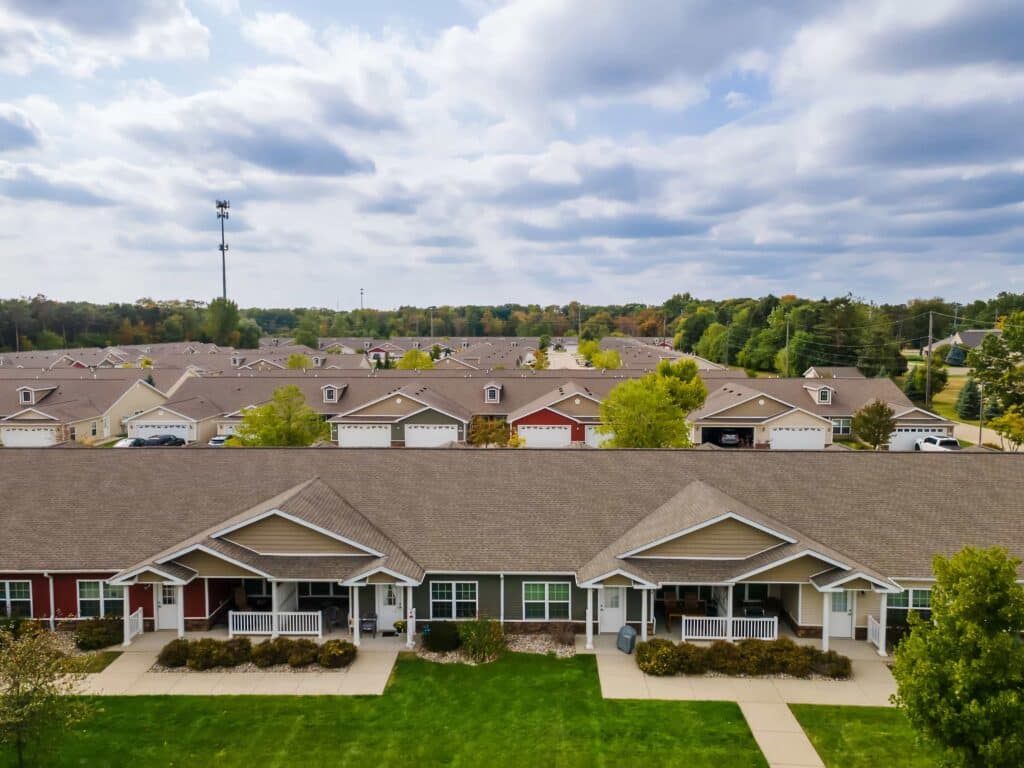 A row of single-story townhouses with porches faces a grassy lawn; more similar homes and trees are visible in the background under a partly cloudy sky.