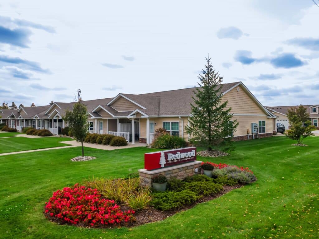Single-story residential buildings with beige siding and white trim, landscaped lawns, and a red sign labeled "Redwood" in the foreground.