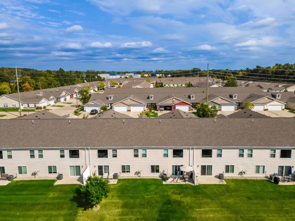 Aerial view of suburban townhouses with green lawns, paved driveways, and a backdrop of trees under a partly cloudy sky.