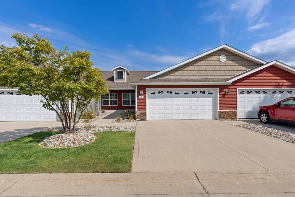 A duplex with a red and beige exterior, two white garage doors, a driveway, a tree, and a parked red car on the right.