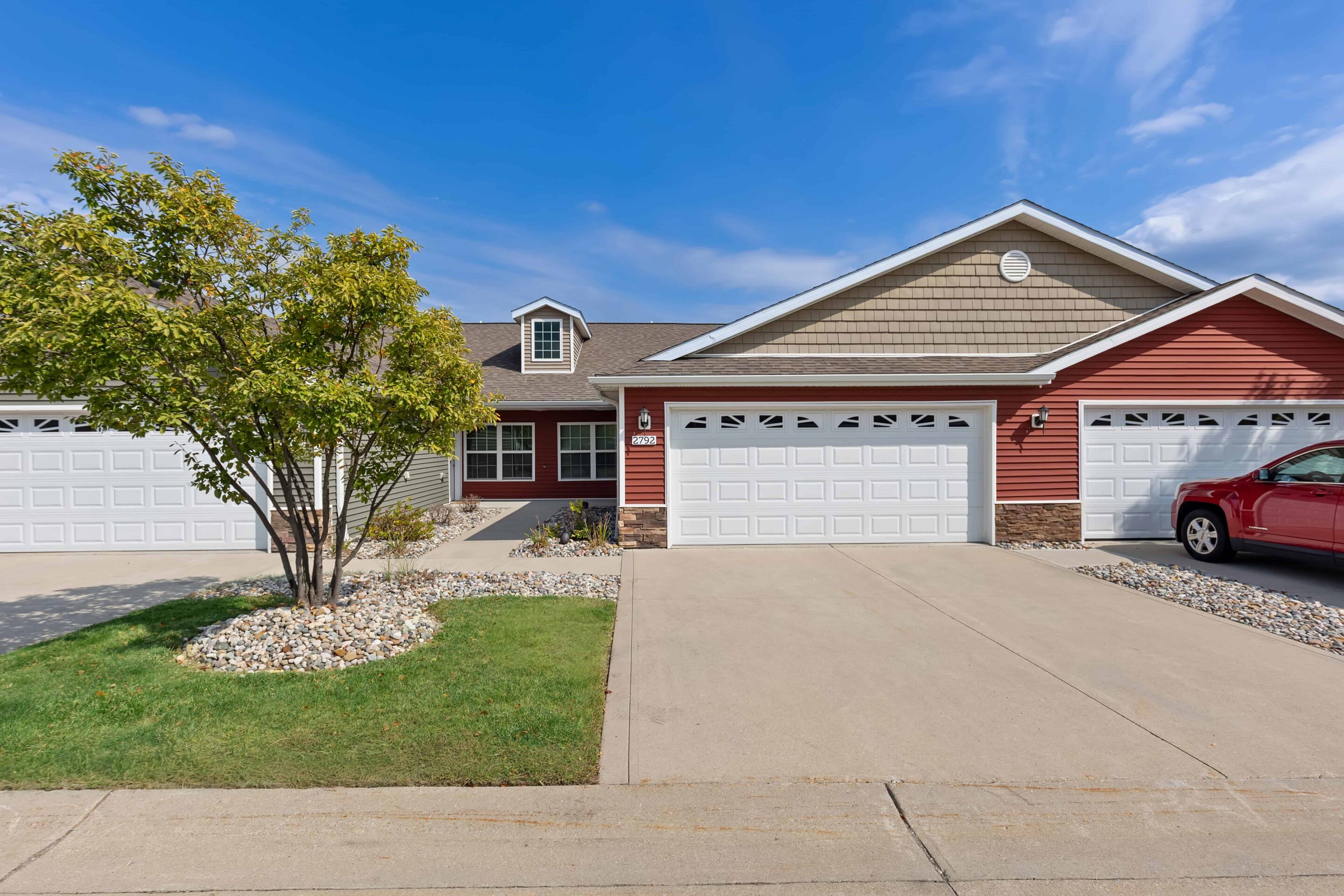 A duplex with a red and beige exterior, two white garage doors, a driveway, a tree, and a parked red car on the right.