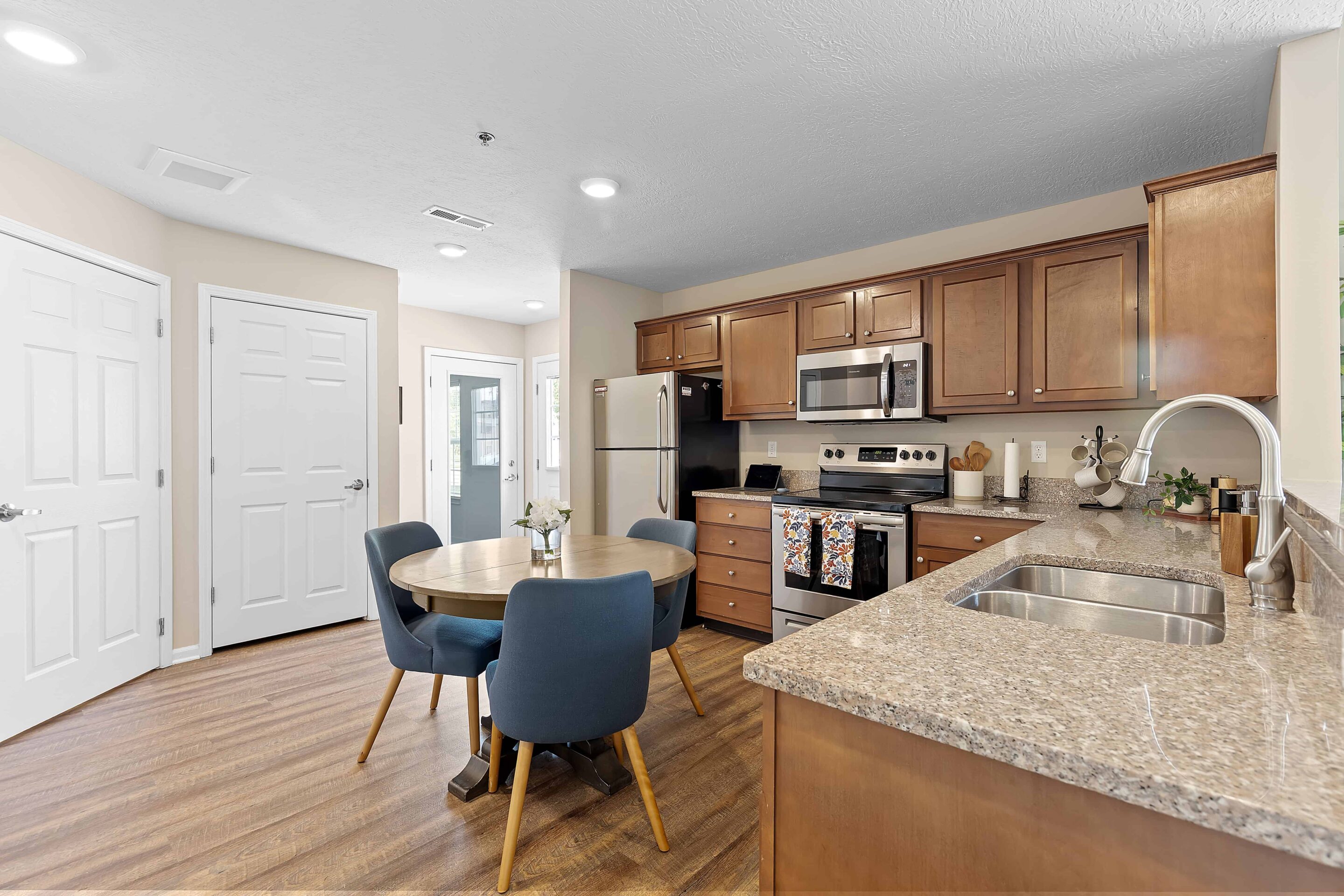 Modern kitchen with brown cabinets, stainless steel appliances, a round dining table with four blue chairs, and wood-style flooring.