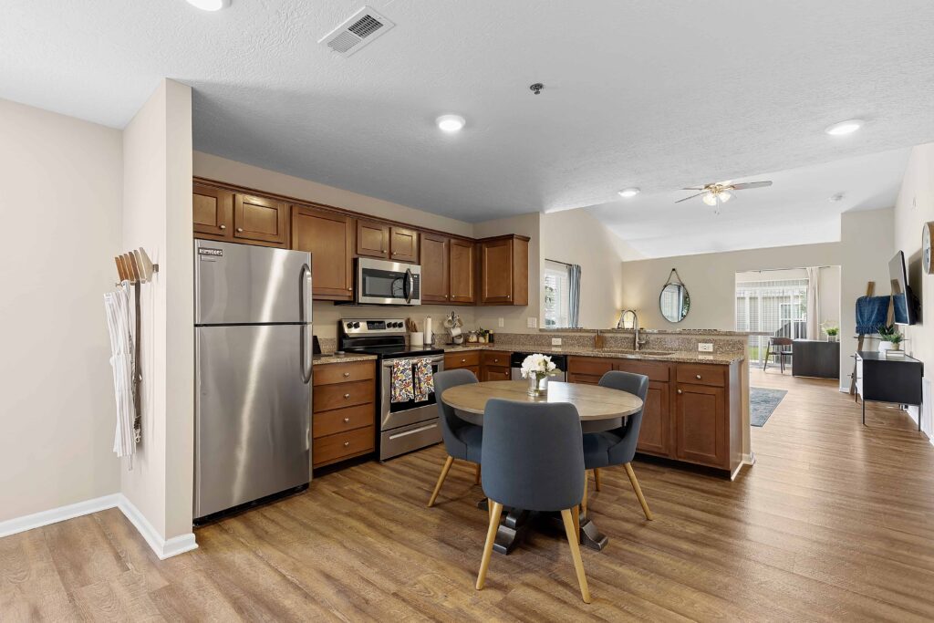 Modern kitchen with stainless steel appliances, wooden cabinets, round dining table with four chairs, and an open layout leading to the living room.