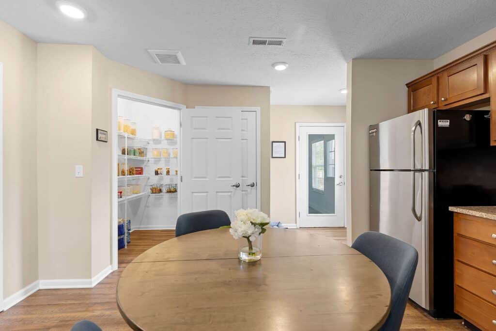 A kitchen with a round wooden dining table, four chairs, stainless steel refrigerator, and an open pantry with shelves stocked with food items.