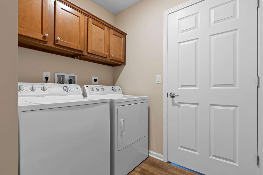 A laundry room with a washing machine and dryer side by side, wooden cabinets above, and a closed white door to the right.