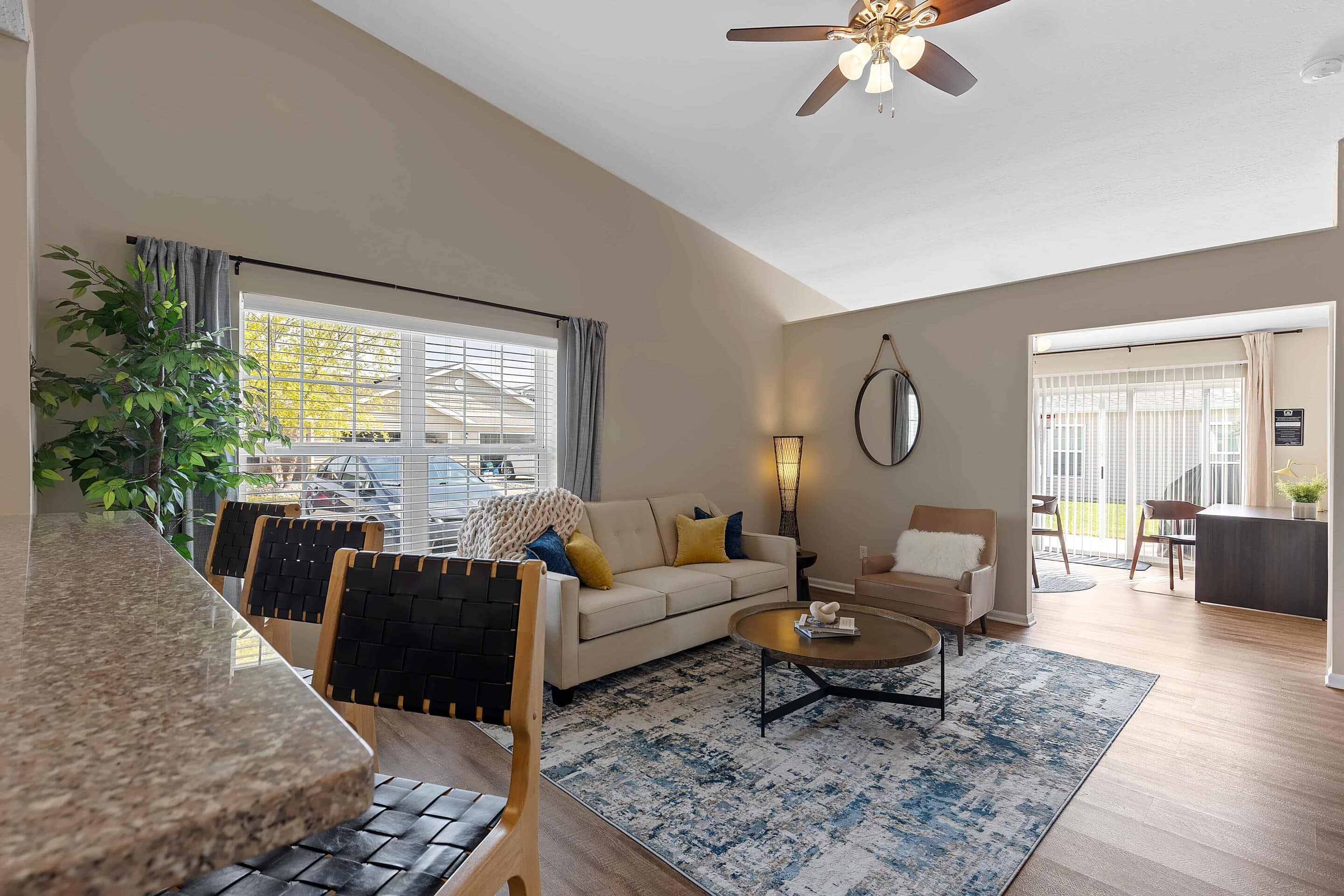 Bright living room with a beige sofa, armchair, rug, round coffee table, and large window; dining area and office space visible in the background.