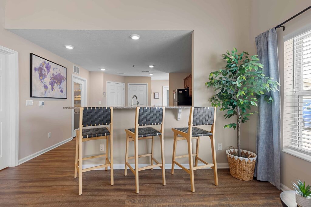Three black and wood barstools are lined up at a kitchen counter in a modern, bright room with wood flooring, wall art, and a potted plant.