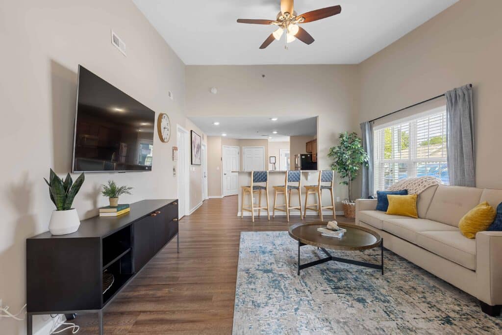 Modern living room with a wall-mounted TV, beige sofa, coffee table, and dining area with four chairs; large window allows natural light into the space.