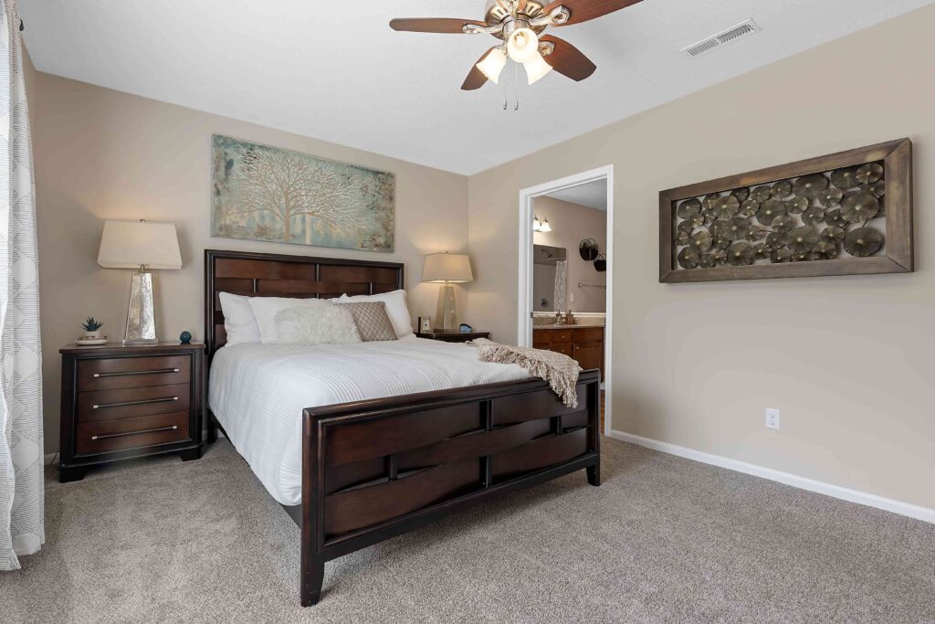 A bedroom with a dark wood bed, two bedside tables with lamps, neutral walls, carpeted floor, ceiling fan, and an open door leading to a bathroom.