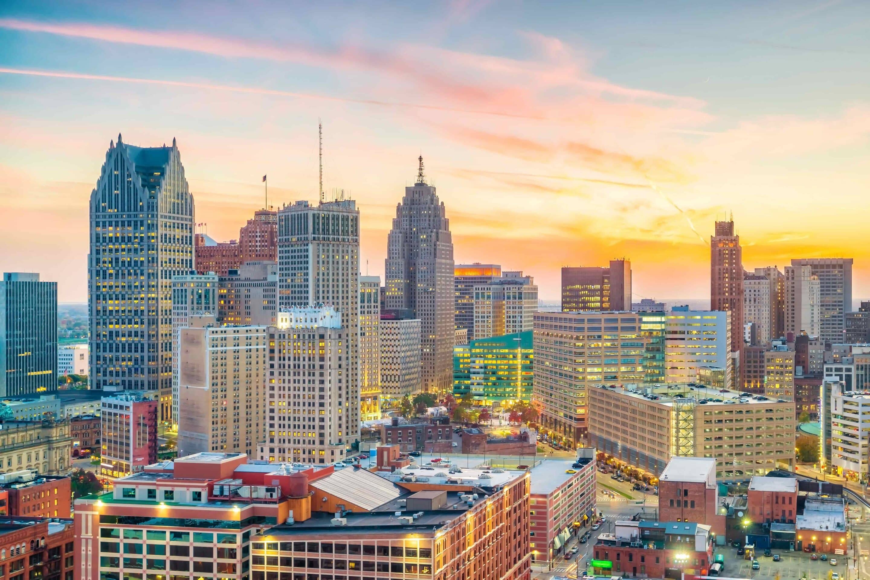 A cityscape view of downtown Detroit at sunset, with tall buildings, offices, and streets visible under a colorful sky.