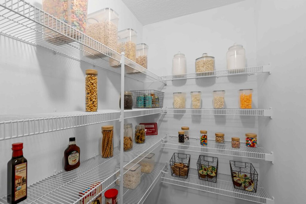 A pantry with white wire shelves holding jars of cereals, pasta, snacks, and condiments, as well as metal baskets containing various food items.