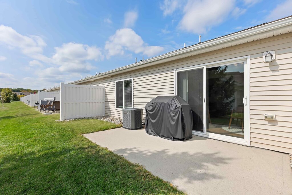 A concrete patio with a covered grill and air conditioning unit beside a beige-sided building with sliding glass doors and a grassy lawn.