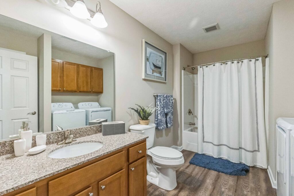 A bathroom with a sink, toilet, and shower with a white curtain; a laundry area with washer and dryer is visible in the background.
