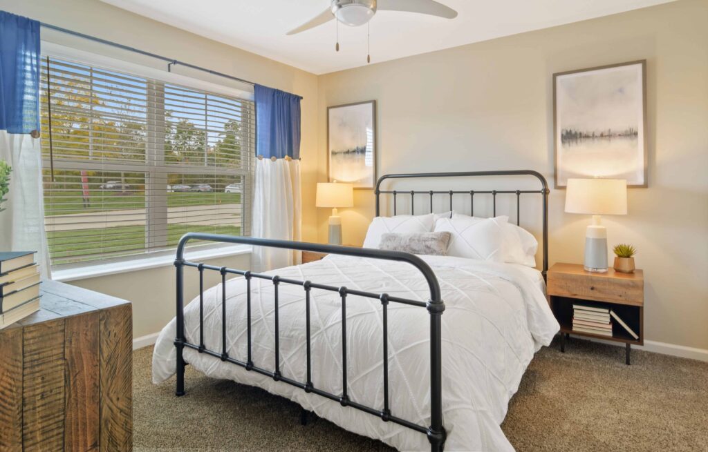 A neatly made bed with a metal frame is centered in a bedroom with beige walls, two nightstands, table lamps, and large window with blue and white curtains.