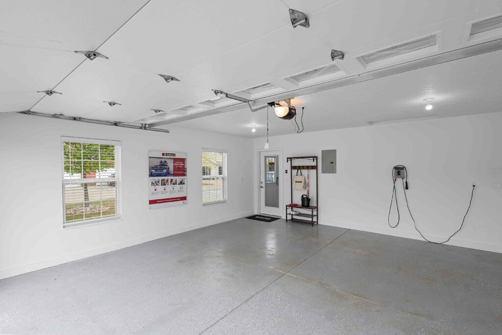 Clean, empty garage with light gray flooring, white walls, wall-mounted electric vehicle charger, a metal shelf, and two windows; a poster and electrical panel are visible on the wall.