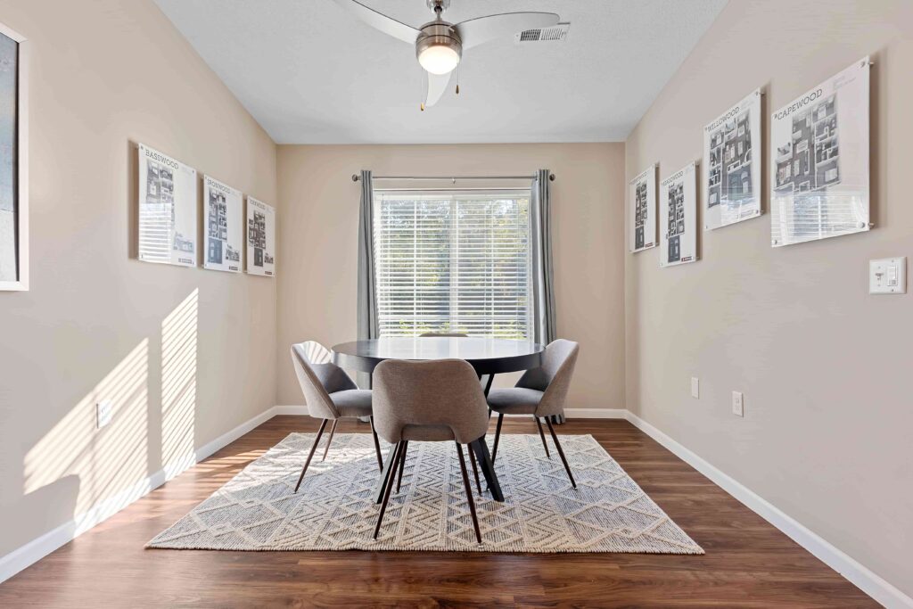 A small dining area with a round black table, four gray chairs, a patterned rug, wall art, and a window with blinds and curtains.