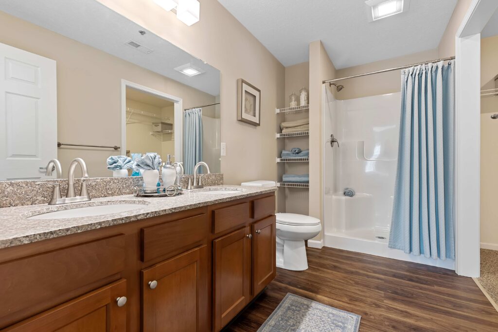 Modern bathroom with dual-sink vanity, granite countertop, wooden cabinets, toilet, walk-in shower with blue curtain, shelving, and wood-look flooring.