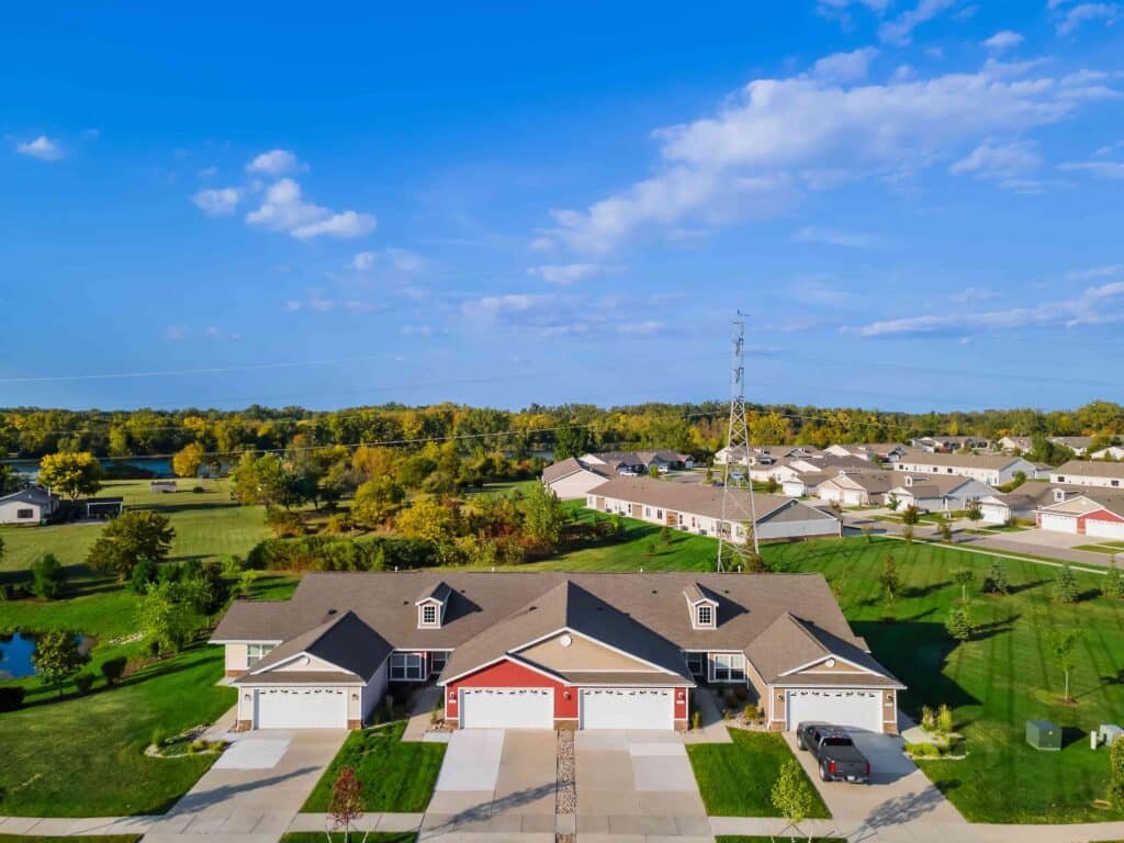 Aerial view of suburban homes with garages, green lawns, scattered trees, and a clear blue sky in the background.