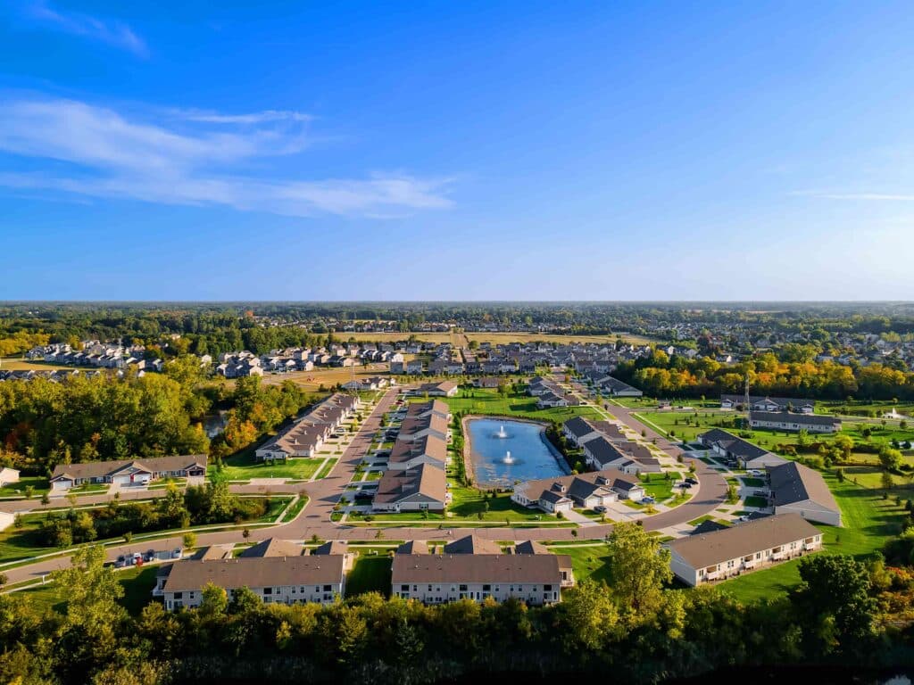 Aerial view of a suburban neighborhood with rows of houses, green lawns, and a central pond under a clear blue sky.