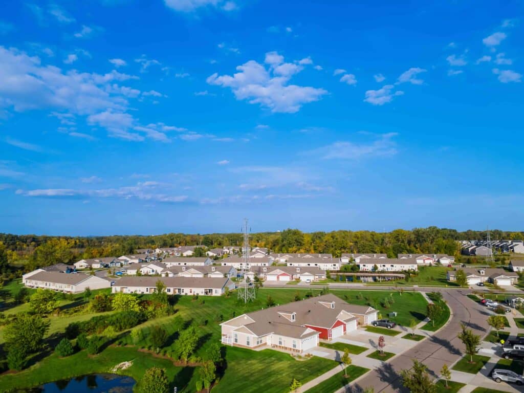 Aerial view of a suburban neighborhood with single-story houses, green lawns, a pond, and a transmission tower under a blue sky with scattered clouds.