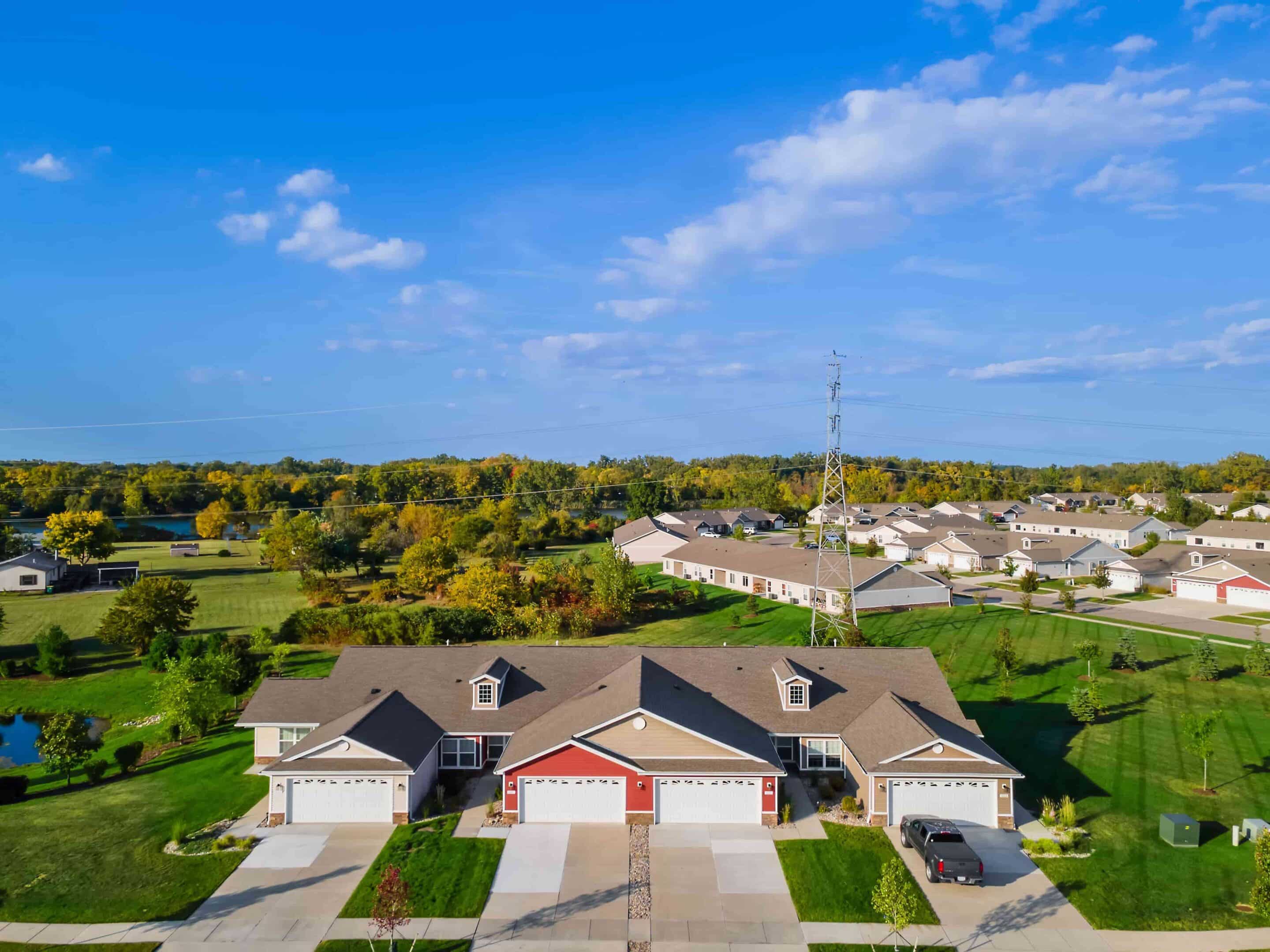 Aerial view of suburban homes with garages, green lawns, scattered trees, and a clear blue sky in the background.