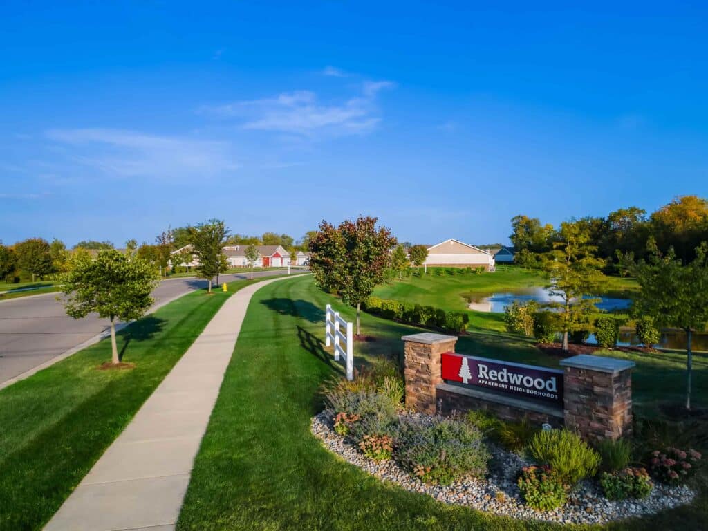 A suburban neighborhood features a sign reading "Redwood Apartment Neighborhoods," a sidewalk, manicured lawns, small trees, and a pond with a fountain under a clear blue sky.