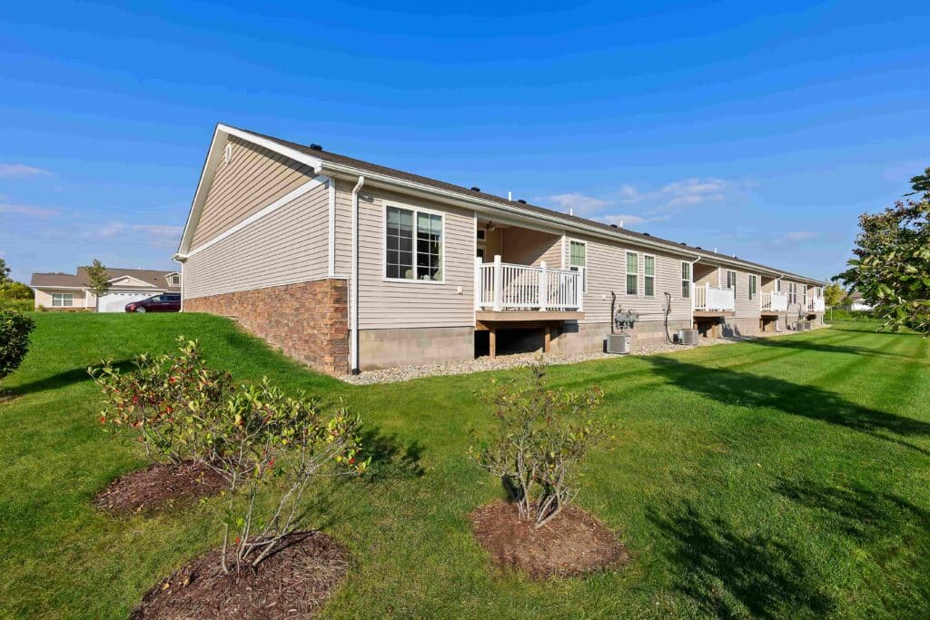 Single-story tan townhomes with small back decks and landscaped lawns on a sunny day.