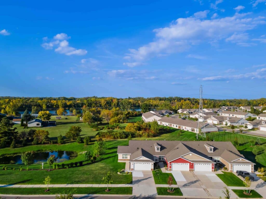 Aerial view of a suburban neighborhood with single-story homes, green lawns, a small pond, trees, and open fields under a blue sky with scattered clouds.