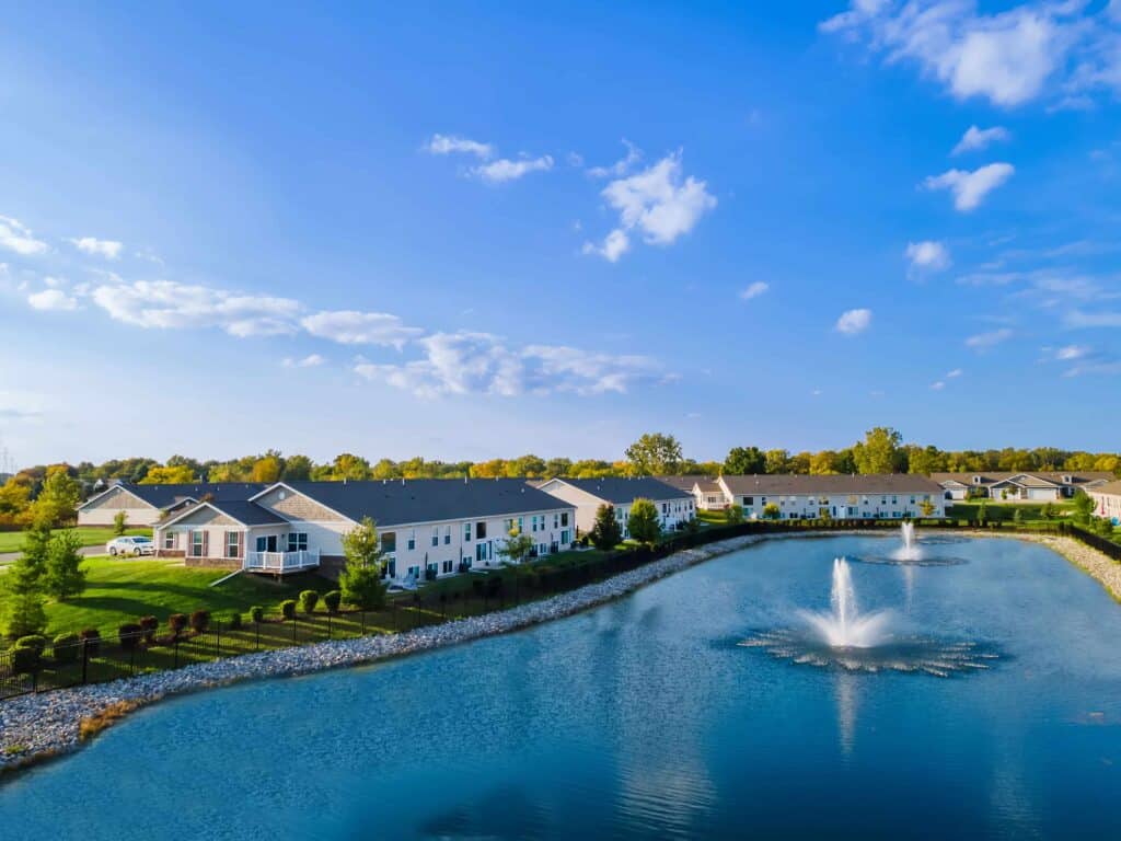 A row of residential homes lines a pond with two water fountains, surrounded by green lawns and trees under a blue sky with scattered clouds.