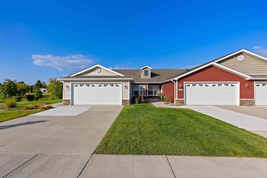 A duplex with red siding, white garage doors, and driveway, surrounded by green grass under a clear blue sky.