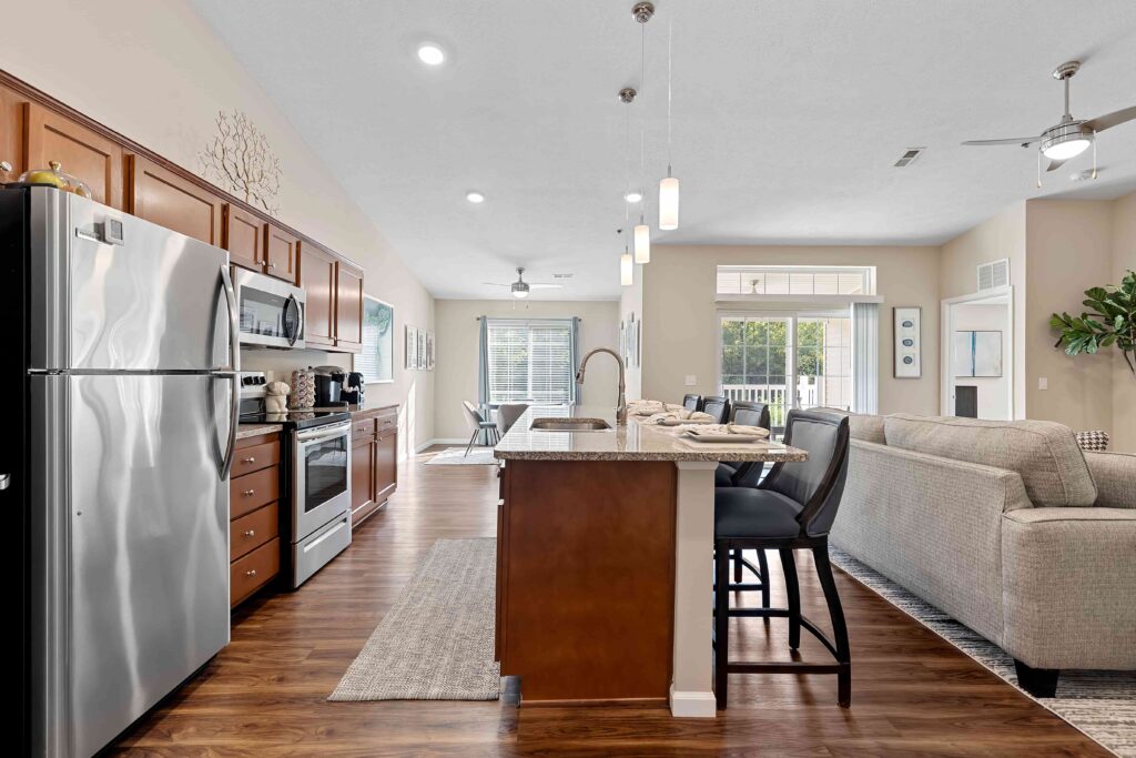 Open-concept kitchen and living room with stainless steel appliances, wooden cabinets, island seating, and a gray sectional sofa. Large windows provide natural light.