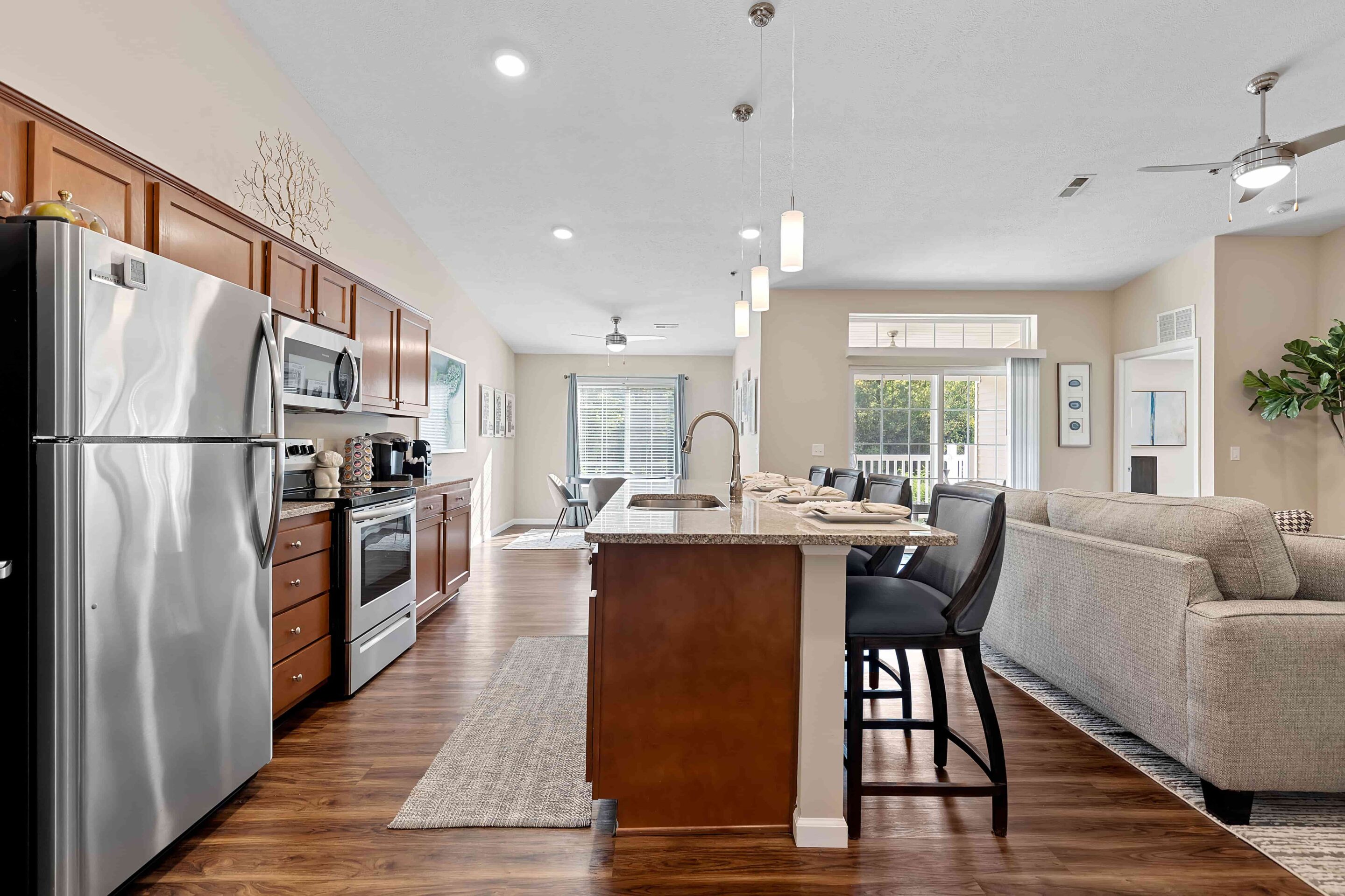 Open-concept kitchen and living room with stainless steel appliances, wooden cabinets, island seating, and a gray sectional sofa. Large windows provide natural light.