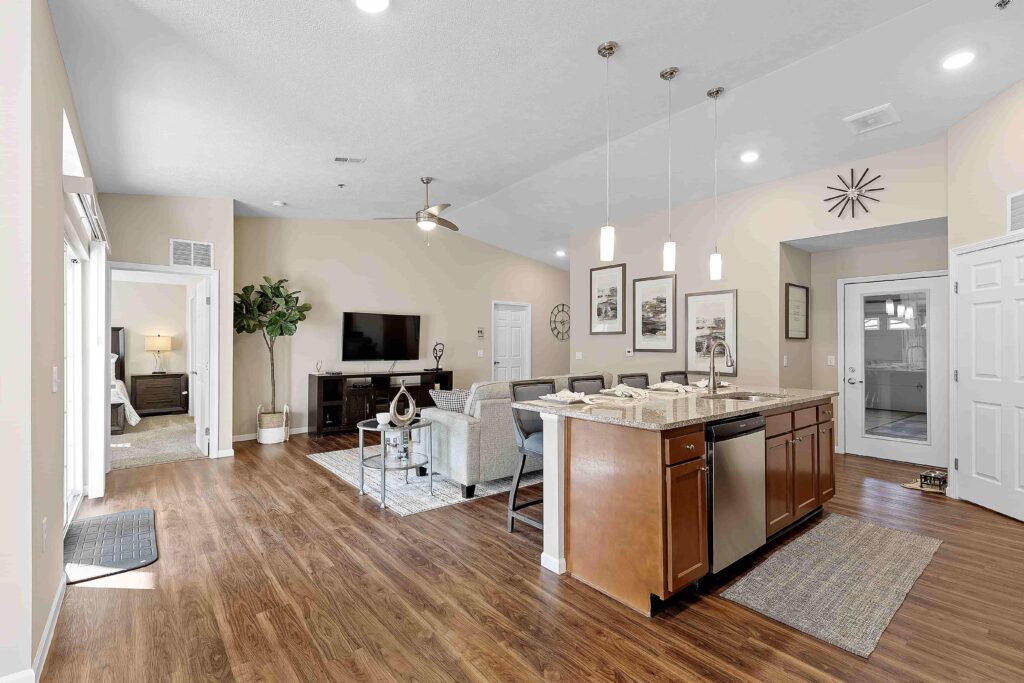Modern open-concept living space featuring a kitchen island, stainless steel appliances, living area with TV, and wood flooring throughout. Bedroom visible through open doorway.