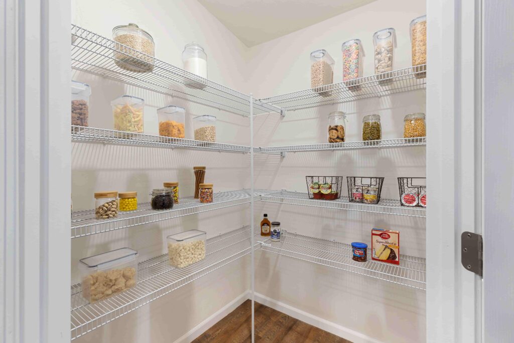 A walk-in pantry with wire shelves holds neatly arranged clear containers of dry goods, jars, baskets with snacks, and canned items on a wooden floor.
