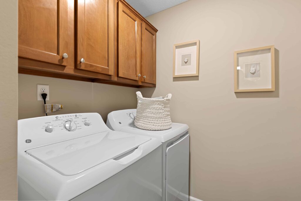 A laundry room with a white washer and dryer, wooden cabinets above, a woven basket on the washer, and two framed artworks on a beige wall.