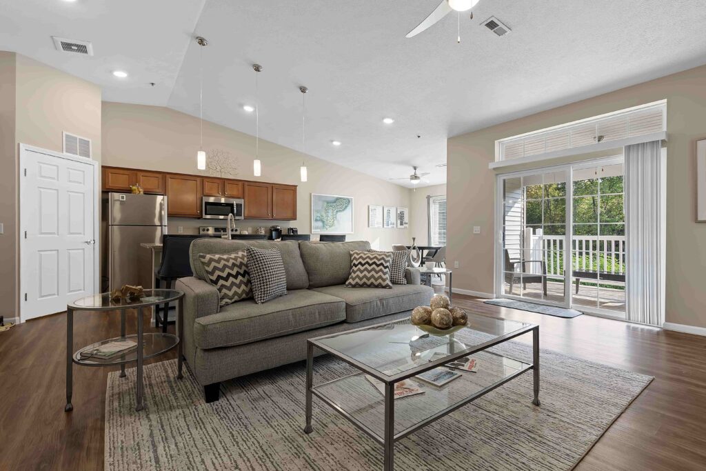 Open-concept living room and kitchen with gray sofa, glass coffee table, stainless steel appliances, and sliding doors leading to a balcony with a green outdoor view.