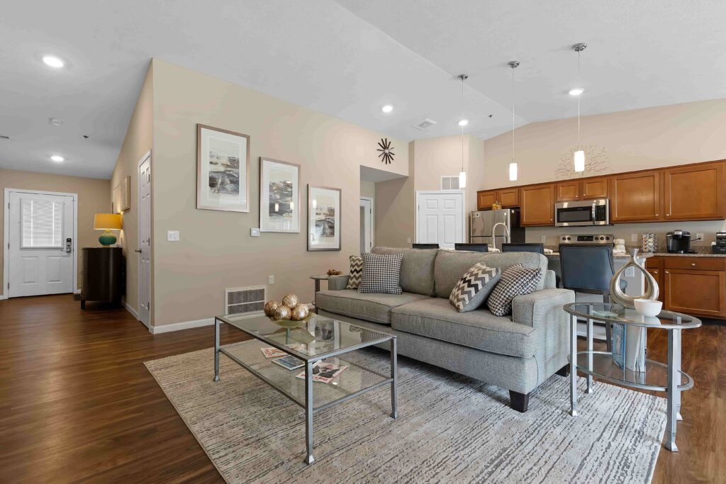 Modern open-concept living room and kitchen with neutral decor, a gray sofa, glass coffee table, wood cabinets, and stainless steel appliances.