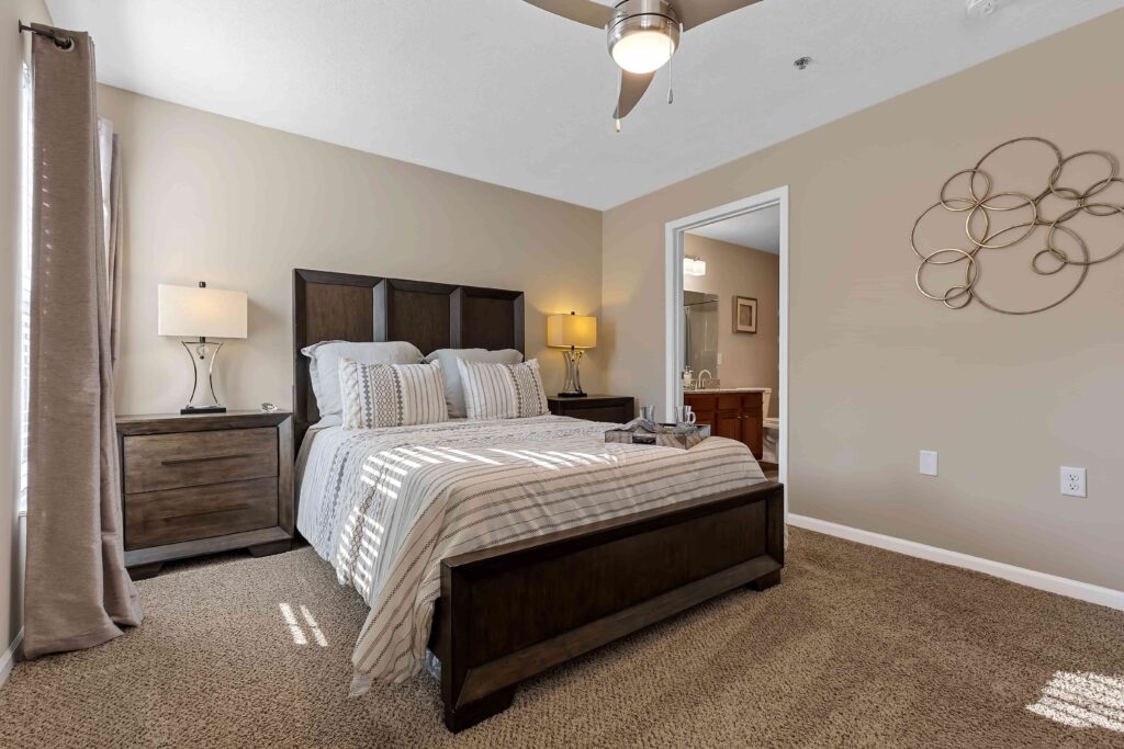 A neatly made bed with striped bedding is centered between two nightstands in a carpeted bedroom with beige walls and a ceiling fan.
