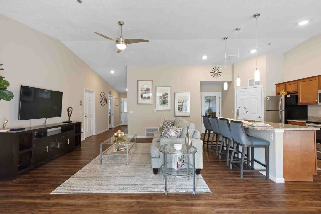 Open-plan living room and kitchen with wood flooring, modern furnishings, a wall-mounted TV, pendant lighting, and a breakfast bar with four barstools.