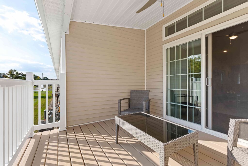 A small balcony with two wicker chairs, a glass-top table, beige siding, white railing, and sliding glass doors.