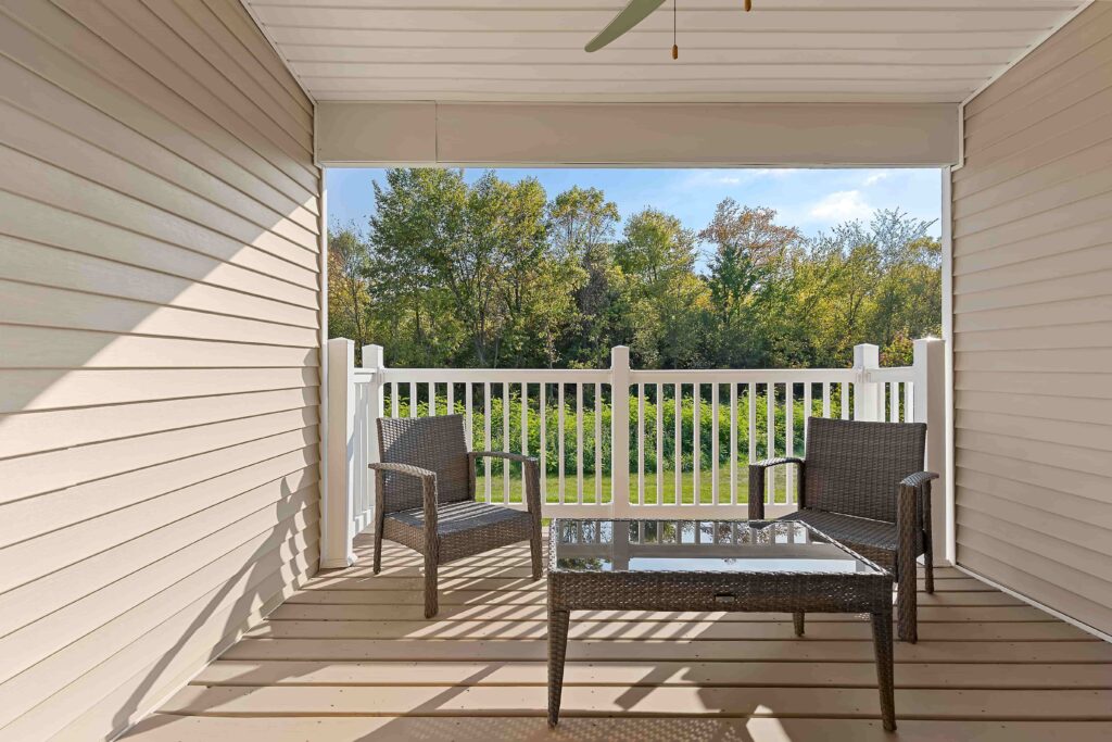 Covered porch with two wicker chairs and a glass-top table, white railing, and a view of trees and greenery in the background.