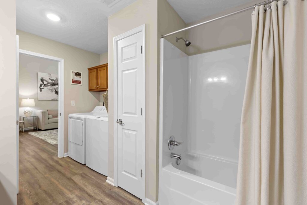 A bathroom with a white bathtub and shower curtain next to a laundry area with a washing machine, dryer, and white cabinets, leading into a living room.