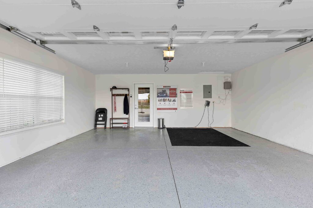 A clean, empty garage with light gray floors, white walls, a window on the left, storage shelves at the back, and an open overhead door.