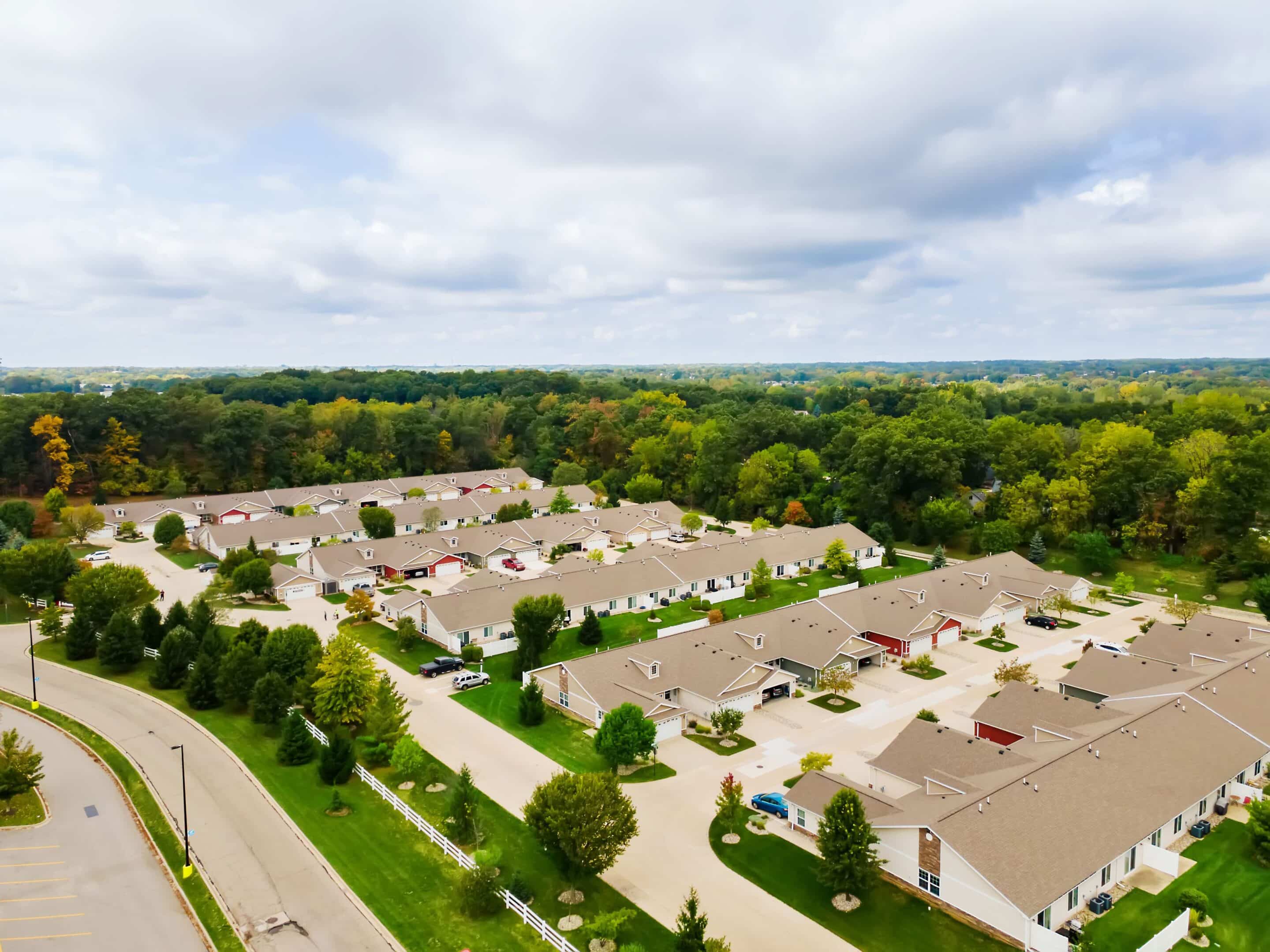 Aerial view of a suburban neighborhood with rows of single-story houses, tree-lined streets, and a forested area in the background under a cloudy sky.