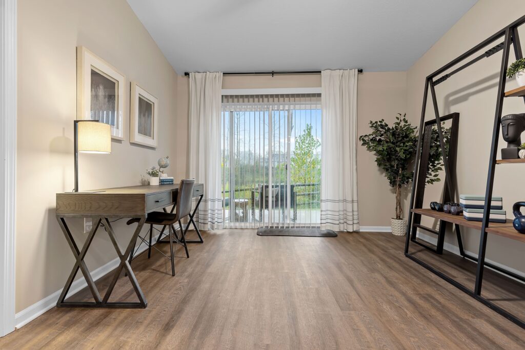 Home office with a desk and chair on the left, open shelving on the right, wood flooring, and large sliding glass doors with white curtains leading to an outdoor view.